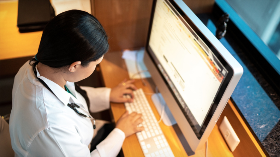 woman in a white lab coat and stethoscope around neck typing at a computer